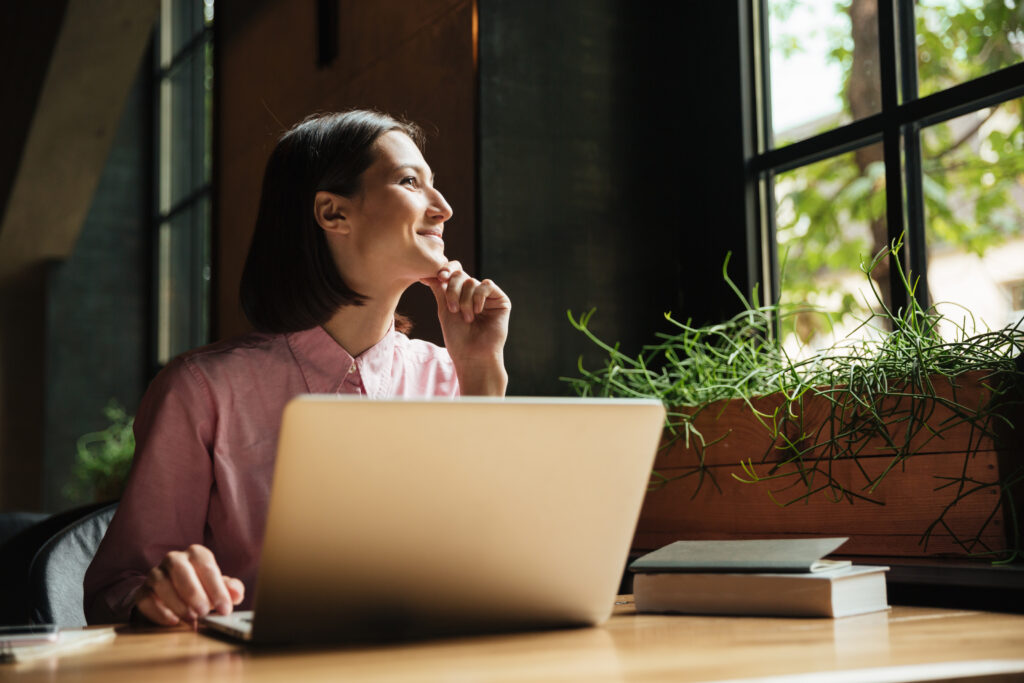 Uma mulher sorridente está sentada em frente a uma janela com seu notebook, enquanto busca oportunidades para sua transição de carreira de TI.