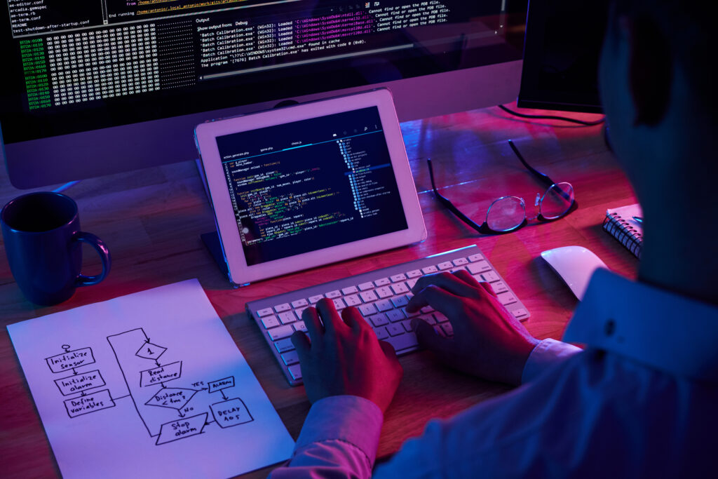Um profissional de desenvolvimento está programando com linguagens de alta e baixa plataforma. Há um profissional de costas sentado em uma mesa com computador, tablet, um rascunho de papel com o projeto desenhado, uma caneca e óculos de grau.