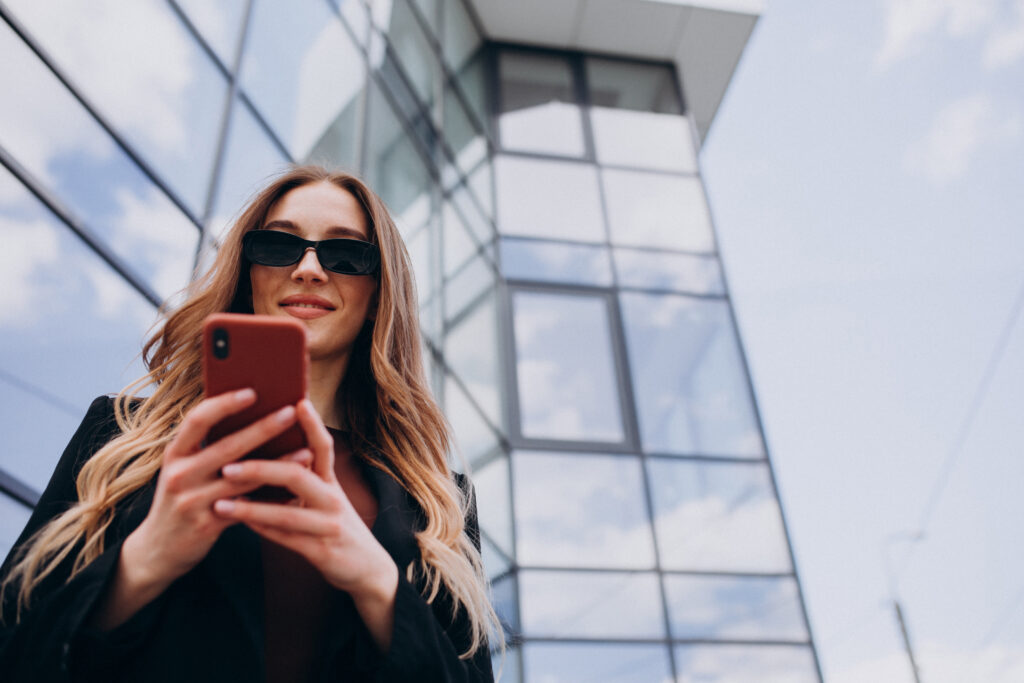 Uma mulher sorridente de óculos escuros está usando um celular em frente a uma torre comercial, representando um profissional outlier em TI.