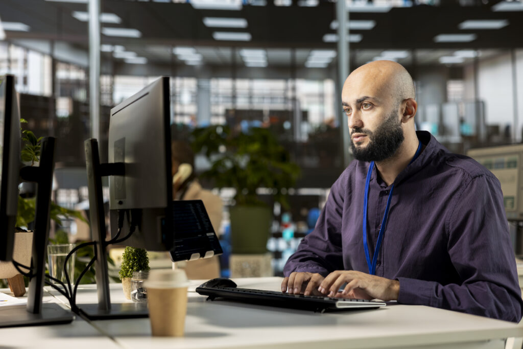 Quais são os profissionais de TI mais difíceis de contratar? 1 Homem, representando os profissionais de TI mais difíceis de contratar, vestindo um moletom, trabalhando em um escritório. Ele está sentado enquanto digita no computador e há um copo de café em sua mesa.