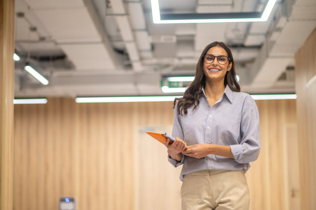 Mulher sorridente de óculos em um ambiente corporativo, representando a carreira de TI.