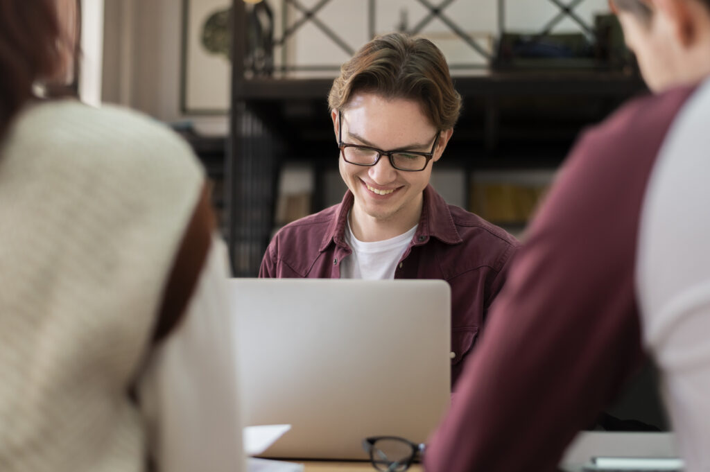 Um homem sorridente de óculos está utilizando um notebook em frente a dois recrutadores que estão tentando contratar desenvolvedor sênior.
