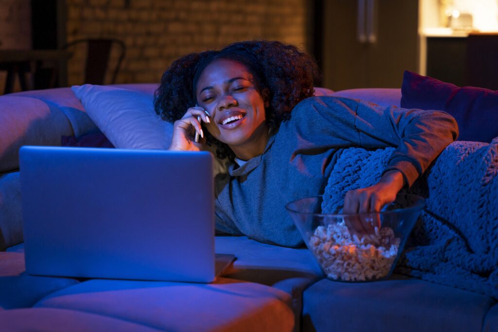 12 séries sobre tecnologia para quem ama o tema 1 Uma mulher sorridente está deitada em um sofá com um balde de pipoca enquanto assiste séries sobre tecnologia no notebook.