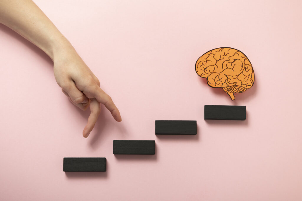 Uma mão gesticula simulando subir uma mini escada. No último degrau há o desenho de um cérebro que representa o agentic mindset.