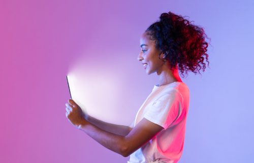 Side profile of happy black woman with curly hair, looking at her tablet whose screen illuminates her face, against purple and pink gradient neon backdrop