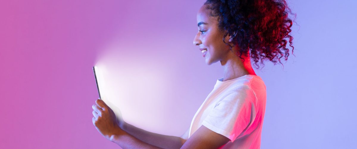 Side profile of happy black woman with curly hair, looking at her tablet whose screen illuminates her face, against purple and pink gradient neon backdrop