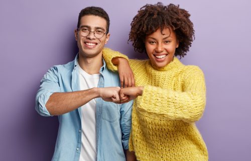Mixed race partners give fist bump, agree to work together, have happy facial expressions rejoice success and cooperation, have toothy smiles, isolated over purple background. Team work concept