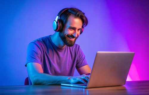 Freelance And Gaming. Happy Millennial Guy Wearing Headphones Browsing Internet On Laptop Computer, Sitting At Desk While Working Or Playing Online Game Over Neon Purple