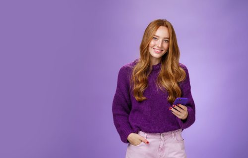 Friendly-looking relaxed and joyful nice 20s female student writing post in social network using mobile phone holding one hand in pocket and texting friend with smartphone, smiling satisfied.