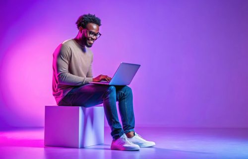 Handsome Black Programmer Guy Using Laptop Computer, Sitting On Big Cube In Neon Lighting, Cheerful African American Man In Glasses Working Or Study Online Over Purple