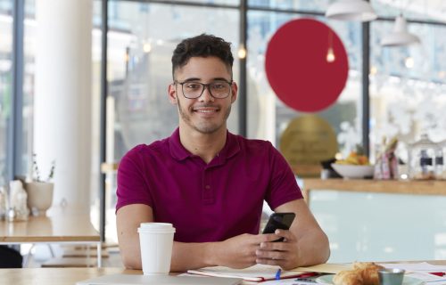 Handsome cheerful young man searches job, browses webpage on modern cell phone, checks information in internet, sits at cafeteria, enjoys sweet dessert and fresh drink, looks gladfully at camera