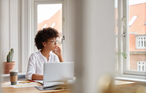 Photo of female entrepreneur speaks via cell phone, looks thoughtfully into window, analyzes paperwork from accounting department, checks database, sit at workplace, disposable coffee, laptop on desk