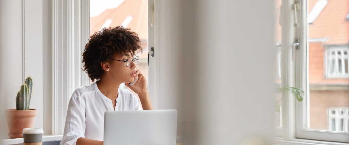 Photo of female entrepreneur speaks via cell phone, looks thoughtfully into window, analyzes paperwork from accounting department, checks database, sit at workplace, disposable coffee, laptop on desk
