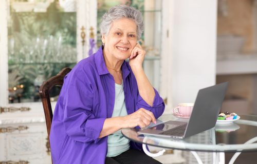Senior Woman Using Laptop Computer And Smiling To Camera Posing At Table In Cozy Cafe Interior, Drinking Coffee And Having Dessert. Elderly Lady At PC Surfing Web On Weekend. Internet Technology