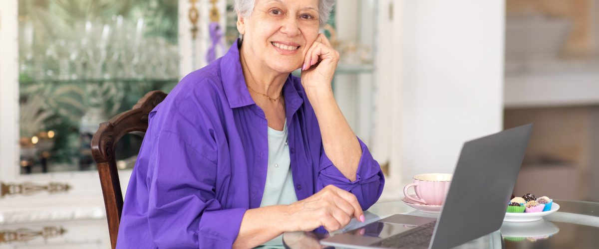 Senior Woman Using Laptop Computer And Smiling To Camera Posing At Table In Cozy Cafe Interior, Drinking Coffee And Having Dessert. Elderly Lady At PC Surfing Web On Weekend. Internet Technology