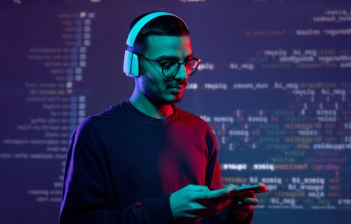 Serious advanced young Arabian tester in wireless headphones examining new app on gadget, coding background