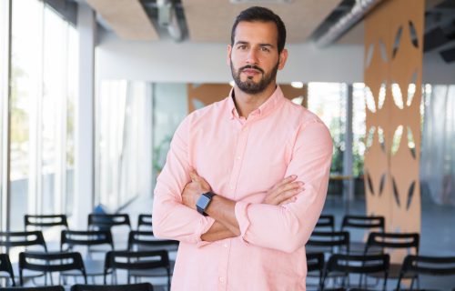 Serious confident business coach posing with arms folded. Young man wearing casual pink shirt, standing in training room and looking at camera. Business training concept