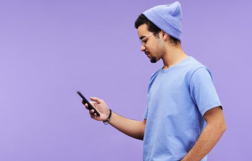 Side view of serious guy in blue t-shirt and beanie hat texting in mobile phone or looking through online information on violet background