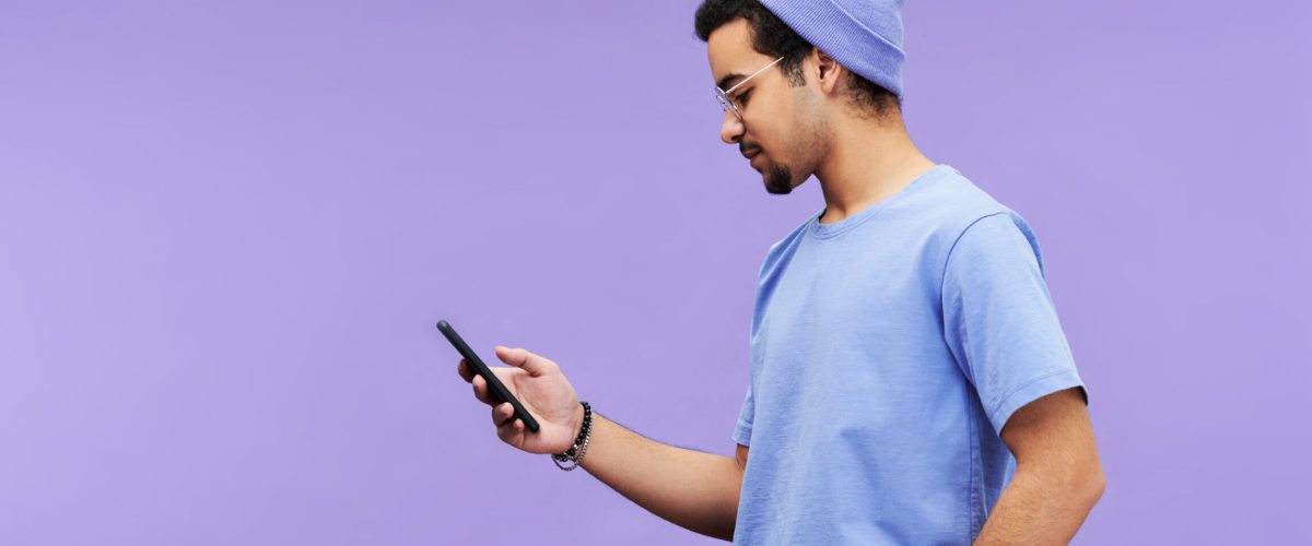 Side view of serious guy in blue t-shirt and beanie hat texting in mobile phone or looking through online information on violet background