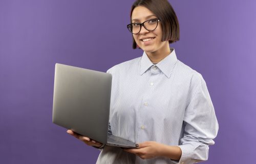 smiling young call center girl wearing glasses holding laptop isolated on purple background