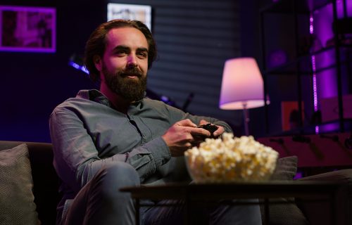 Cheerful gamer sitting on couch engaging in online competitive multiplayer tourney using gaming console and gamepad. Upbeat player engaging in fun videogames, enjoying leisure time, camera A