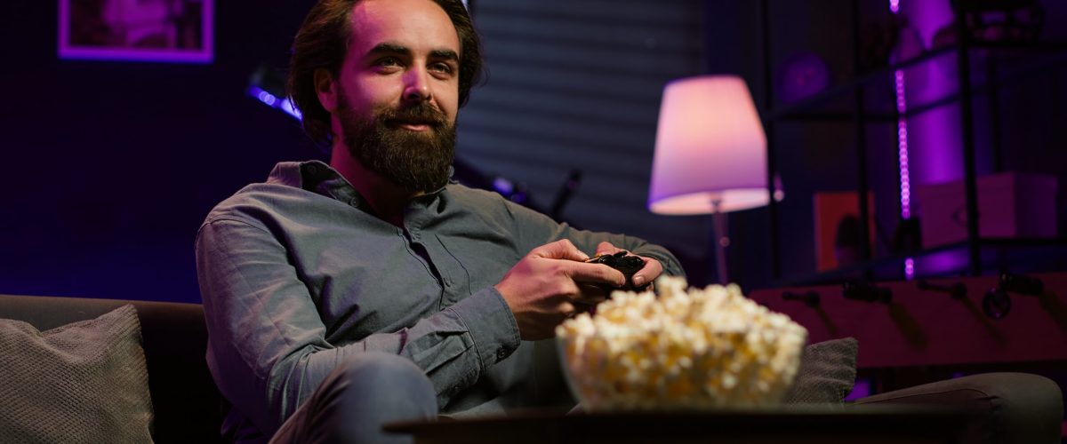Cheerful gamer sitting on couch engaging in online competitive multiplayer tourney using gaming console and gamepad. Upbeat player engaging in fun videogames, enjoying leisure time, camera A